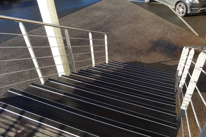 Bilden visar en trappa som leder ner till en parkering. Varje trappsteg är försedd med en trappnos av aluminium (för utomhusbruk) med svart inlägg i profilen. Inlägget/tejpen är av mineral granulat med kornig yta för bästa grepp.